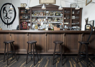 Interiér baru AnonymouS, dřevěný bar, tmavé dřevo, bar jako knihovna, barové židle, logo namalované na zdi