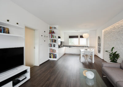 Panelákový byt, obytný prostor, obývák, vinylová podlaha, TV nábytek, konferenční stůl, jídelní stůl, židle, světlo květina, kuchyň, kuchyňská linka, posuvné dveře, knihovna, kamenný obklad