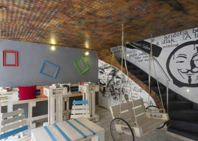 Interiér kavárny AnonymouS, grafika na zdi, dřevěný nábytek, modré pruhy, rezavý strop, houpačka s koly, barevné rámy, logo na zdi