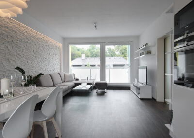 Panelákový byt, obytný prostor, obývák, vinylová podlaha, TV nábytek, konferenční stůl, jídelní stůl, židle, světlo květina, kuchyň, kuchyňská linka, posuvné dveře, knihovna, kamenný obklad, velké balkonové okno