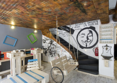 Interiér kavárny AnonymouS, grafika na zdi, dřevěný nábytek, modré pruhy, rezavý strop, houpačka s koly, nástěnné litinové umyvadlo, barevné rámy, logo na zdi, europalety