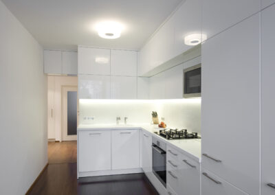 Interiér panelákového bytu, kuchyň, kuchyňská linka, bílá kuchyň, dřevěná podlaha, skříňky