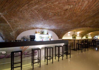 Interiér baru v Poděbradech, cihlové klenby, světlá litá podlaha, tmavé dřevo, barové židle, barový pult, osvětlené police za barem