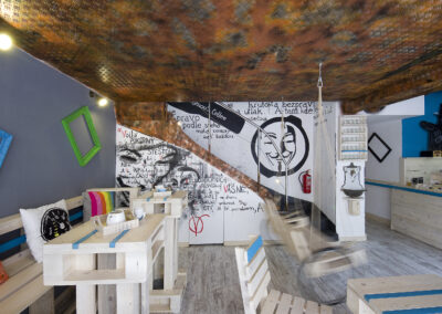 Interiér kavárny AnonymouS, grafika na zdi, dřevěný nábytek, modré pruhy, rezavý strop, houpačka s koly, nástěnné litinové umyvadlo, barevné rámy, logo na zdi