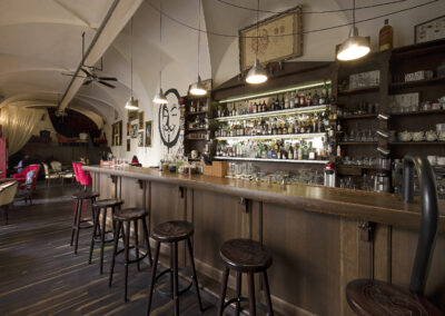 Interiér baru AnonymouS, dřevěný bar, tmavé dřevo, bar jako knihovna, barové židle, logo namalované na zdi, světla nad barem