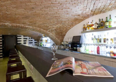 Interiér baru v Poděbradech, cihlové klenby, světlá litá podlaha, tmavé dřevo, barové židle, barový pult, osvětlené police za barem