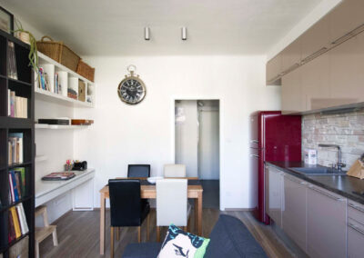 Interiér cihlového bytu, obytný prostor s kuchyní, tmavě šedá pohovka, vinylová podlaha, knihovna, pracovní kout, jídelní stůl, jídelní židle, lanýžová kuchyně