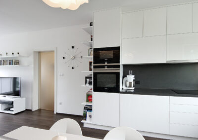 Panelákový byt, obytný prostor, obývák, vinylová podlaha, jídelní stůl, židle, světlo květina, kuchyň, kuchyňská linka, posuvné dveře, knihovna, nástěnné hodiny