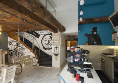 Interiér kavárny AnonymouS, grafika na zdi, dřevěný nábytek, modré pruhy, rezavý strop, houpačka s koly, nástěnné litinové umyvadlo, barevné rámy, modrá zeď, umělecká malba