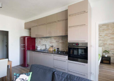 Interiér cihlového bytu, obytný prostor s kuchyní, lanýžová kuchyně, červená retro lednice, kamenný obklad v kuchyni, tmavě šedá pohovka, vinylová podlaha