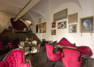 Interiér baru AnonymouS, čalouněné sezení, červená lenoška, tmavé dřevo, schodiště v pozadí, závěs v pozadí, gotické klenby, dřevěné pódium, obrazy na zdi, repliky starožitného nábytku