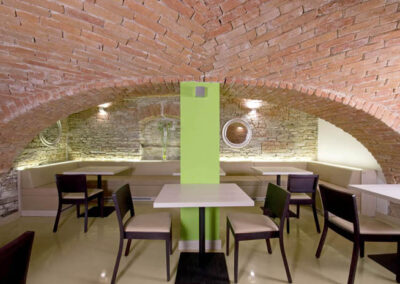 Interiér baru v Poděbradech, cihlové klenby, světlá litá podlaha, tmavé dřevo, jídelní židle, jídelní stoly, zeď ze světlého kamene, zelený sloup