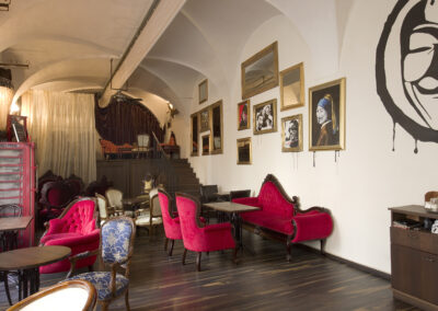 Interiér baru AnonymouS, čalouněné sezení, červená lenoška, tmavé dřevo, schodiště v pozadí, závěs v pozadí, gotické klenby, dřevěné pódium, obrazy na zdi, repliky starožitného nábytku