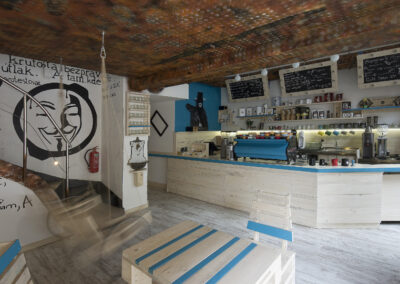 Interiér kavárny AnonymouS, grafika na zdi, dřevěný nábytek, modré pruhy, rezavý strop, houpačka s koly, nástěnné litinové umyvadlo, barevné rámy, modrá zeď, umělecká malba