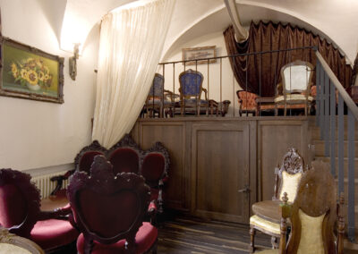 Interiér baru AnonymouS, čalouněné sezení, tmavé dřevo, zlaté čalounění, schodiště v pozadí, závěs v pozadí, gotické klenby, dřevěné pódium, obrazy na zdi, repliky starožitného nábytku