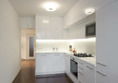 Interiér panelákového bytu, kuchyň, kuchyňská linka, bílá kuchyň, dřevěná podlaha, skříňky