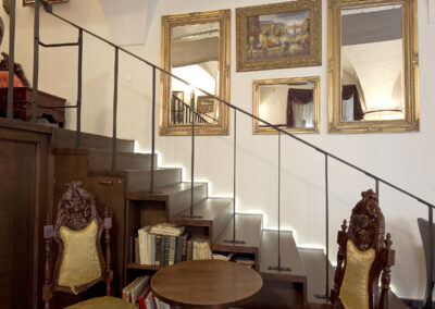 Interiér baru AnonymouS, čalouněné sezení, tmavé dřevo, zlaté čalounění, osvětlené schodiště, závěs v pozadí, gotické klenby, dřevěné pódium, obrazy na zdi, zrcadla na zdi, repliky starožitného nábytku