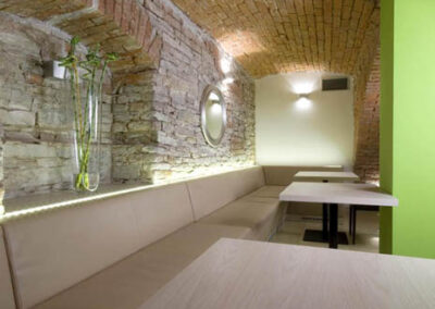 Interiér baru v Poděbradech, cihlové klenby, světlé čalounění, čalouněná lavice, jídelní stoly, zeď ze světlého kamene, zelený sloup, nika v kamenné zdi