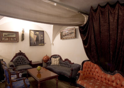 Interiér baru AnonymouS, čalouněné sezení, červená lenoška, tmavé dřevo, závěs v pozadí, gotické klenby, dřevěné pódium, obrazy na zdi, repliky starožitného nábytku, lounge sezení