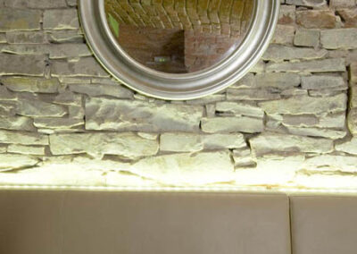 Interiér baru v Poděbradech, světlé čalounění, čalouněná lavice, jídelní stůl, zeď ze světlého kamene, nika v kamenné zdi, nepřímé osvětlení, osvětlení kamenné zdi