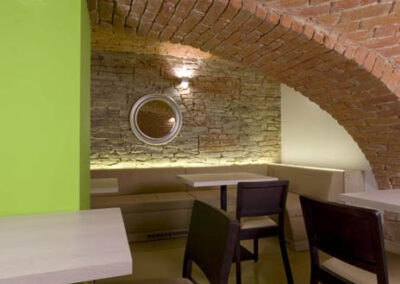 Interiér baru v Poděbradech, světlé čalounění, čalouněná lavice, jídelní stůl, zeď ze světlého kamene, nika v kamenné zdi, nepřímé osvětlení, osvětlení kamenné zdi, zelený sloup, tmavé židle, zrcadlo na zdi