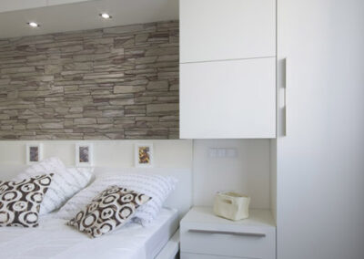 Interiér bytu Řešovská, panelákový byt, kompletní rekonstrukce, ložnice, světlá ložnice, skříně okolo postele, noční stolky, kamenný obklad za postelí, béžový kámen na zdi, osvětlení postele