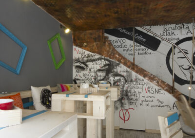 Interiér kavárny AnonymouS, grafika na zdi, dřevěný nábytek, modré pruhy, rezavý strop, barevné rámy na zdi, logo na zdi, sezení u vyšších stolků