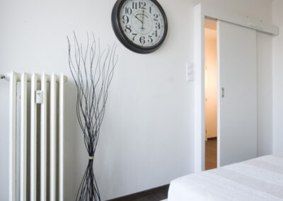 Interiér bytu Řešovská, panelákový byt, kompletní rekonstrukce, ložnice, světlá ložnice, posuvné dveře, hodiny na zdi, retro radiátor