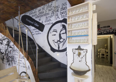 Interiér kavárny AnonymouS, grafika na zdi, dřevěný nábytek, modré pruhy, rezavý strop, houpačka s koly, nástěnné litinové umyvadlo, barevné rámy, logo na zdi, schodiště, nápisy na zdi