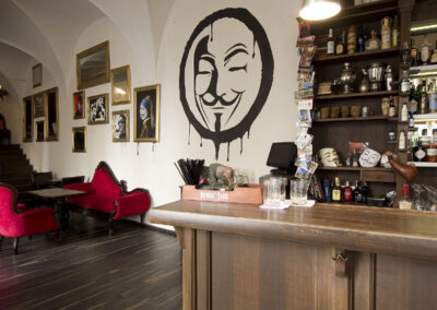 Interiér baru AnonymouS, čalouněné sezení, červená lenoška, tmavé dřevo, obraz na zdi, logo na zdi, bar, vyryté logo na barové židli