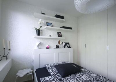 Interiér panelákového bytu, ložnice, bílé police na zdi, závěsné světlo, krémová šatní skříň, tapeta s odlesky