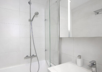 Interiér panelákového bytu, nová koupelna, bílý obklad, zrcadlo, umyvadlo se skříňkou, vana se sprchovou zástěnou, sprchový sloup