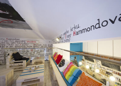 Interiér kavárny AnonymouS, grafika na zdi, dřevěný nábytek, modré pruhy, barevné polštáře, malba na stropě, sezení na dřevěném nábytku, dřevěná lavice, europalety