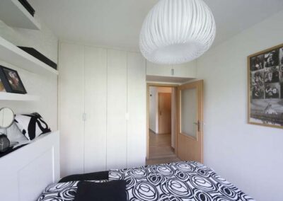 Interiér panelákového bytu, ložnice, bílé police na zdi, závěsné světlo, krémová šatní skříň, pohled do chodby, skříň nade dveřmi