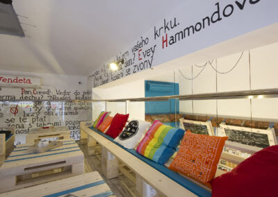 Interiér kavárny AnonymouS, grafika na zdi, dřevěný nábytek, modré pruhy, barevné polštáře, malba na stropě, sezení na dřevěném nábytku, dřevěná lavice, europalety