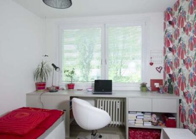 Interiér panelákového bytu, pokoj pro slečnu, pohled na okno, pracovní stůl pod oknem, černé závěsné světlo, postel, červeno bílá tapeta