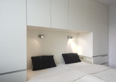 Interiér panelákového bytu, ložnice, skříně okolo postele, postel, šuplíky, osvětlení nad postelí