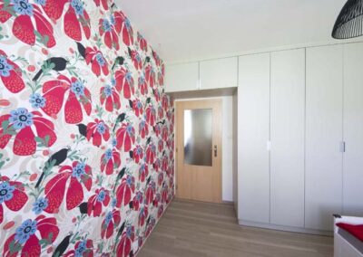 Interiér panelákového bytu, pokoj pro slečnu, černé závěsné světlo, krémová šatní skříň, pohled na dveře, skříň nade dveřmi, červeno bílá tapeta