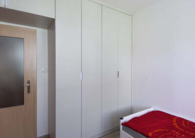 Interiér panelákového bytu, pokoj pro slečnu, černé závěsné světlo, krémová šatní skříň, pohled na dveře, skříň nade dveřmi