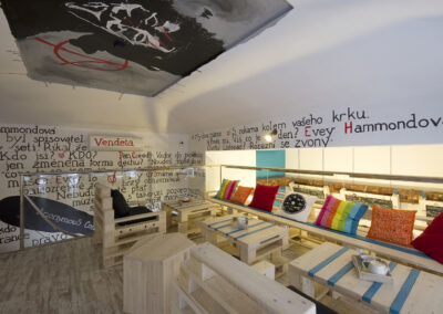 Interiér kavárny AnonymouS, grafika na zdi, dřevěný nábytek, modré pruhy, barevné polštáře, malba na stropě, sezení na dřevěném nábytku, dřevěná lavice, europalety